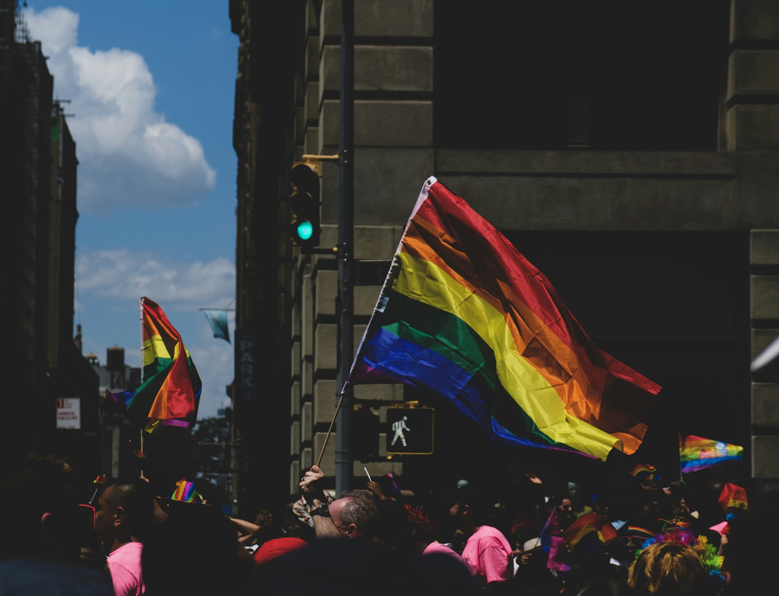 A rainbow flag flying on a city street beside a traffic light.