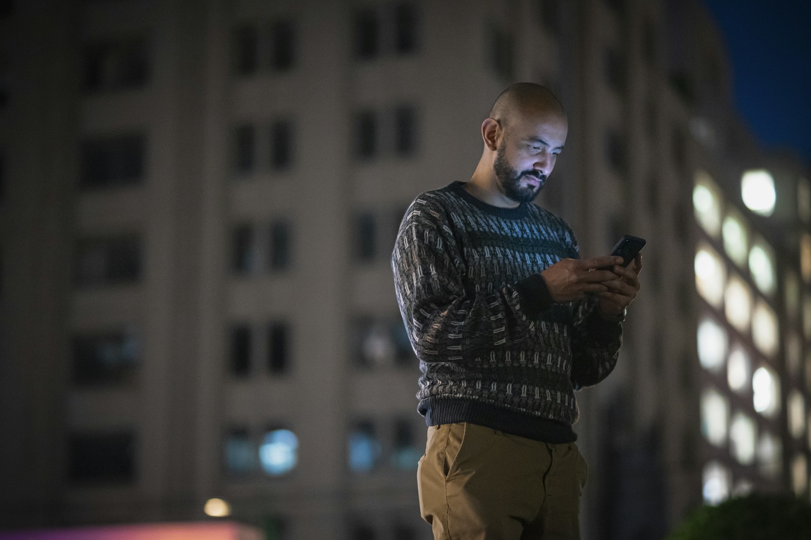 A man standing on a city street at night, lit by his phone screen, tall apartment building behind him.