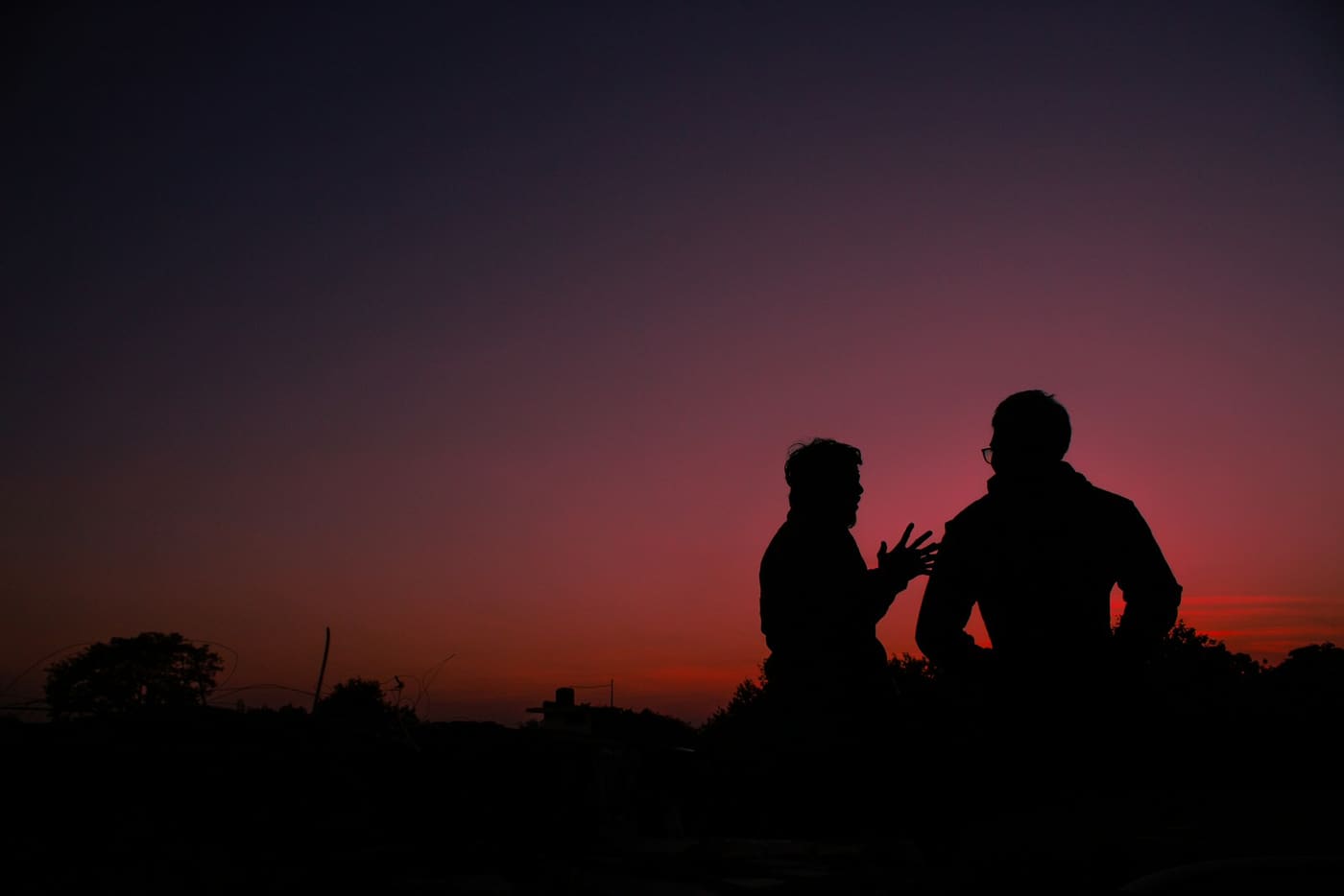 Two men sitting and talking in silhouette against a pink and orange sunset sky.
