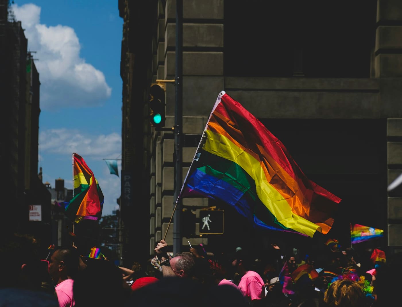A rainbow flag flying on a city street beside a traffic light.