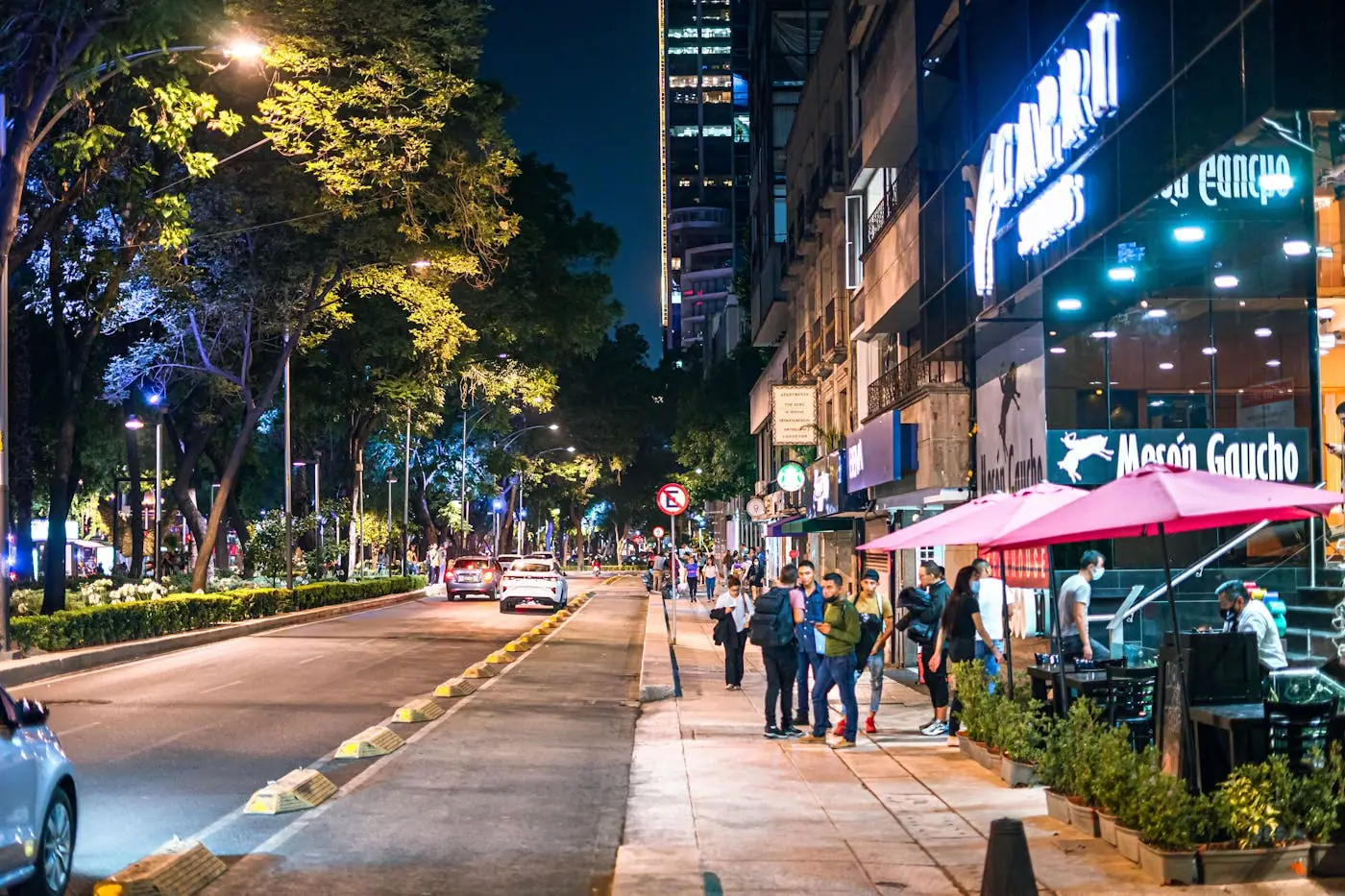 People walking past restaurants and bright lights on a Mexico City street at night.