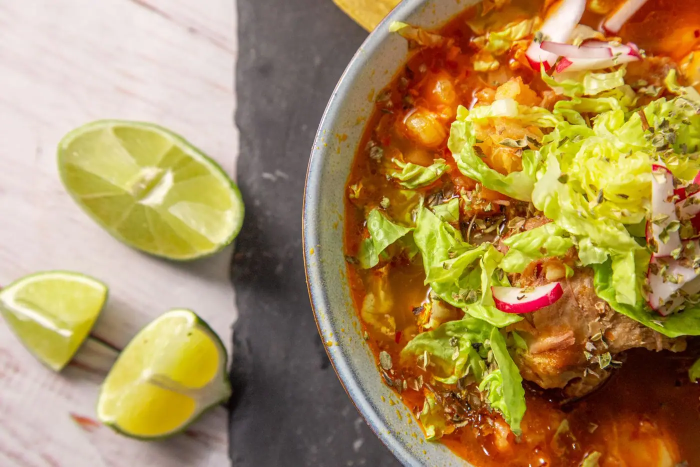 A steaming bowl of red pozole topped with shredded lettuce, radish, and lime on the side.