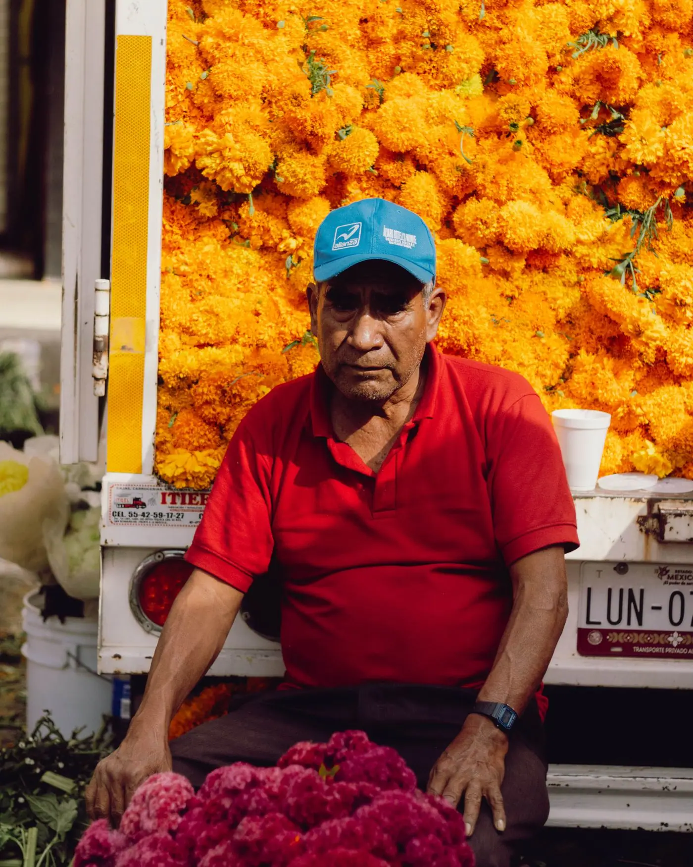 A flower vendor in a red shirt resting by a truck piled high with orange cempasúchil marigolds.