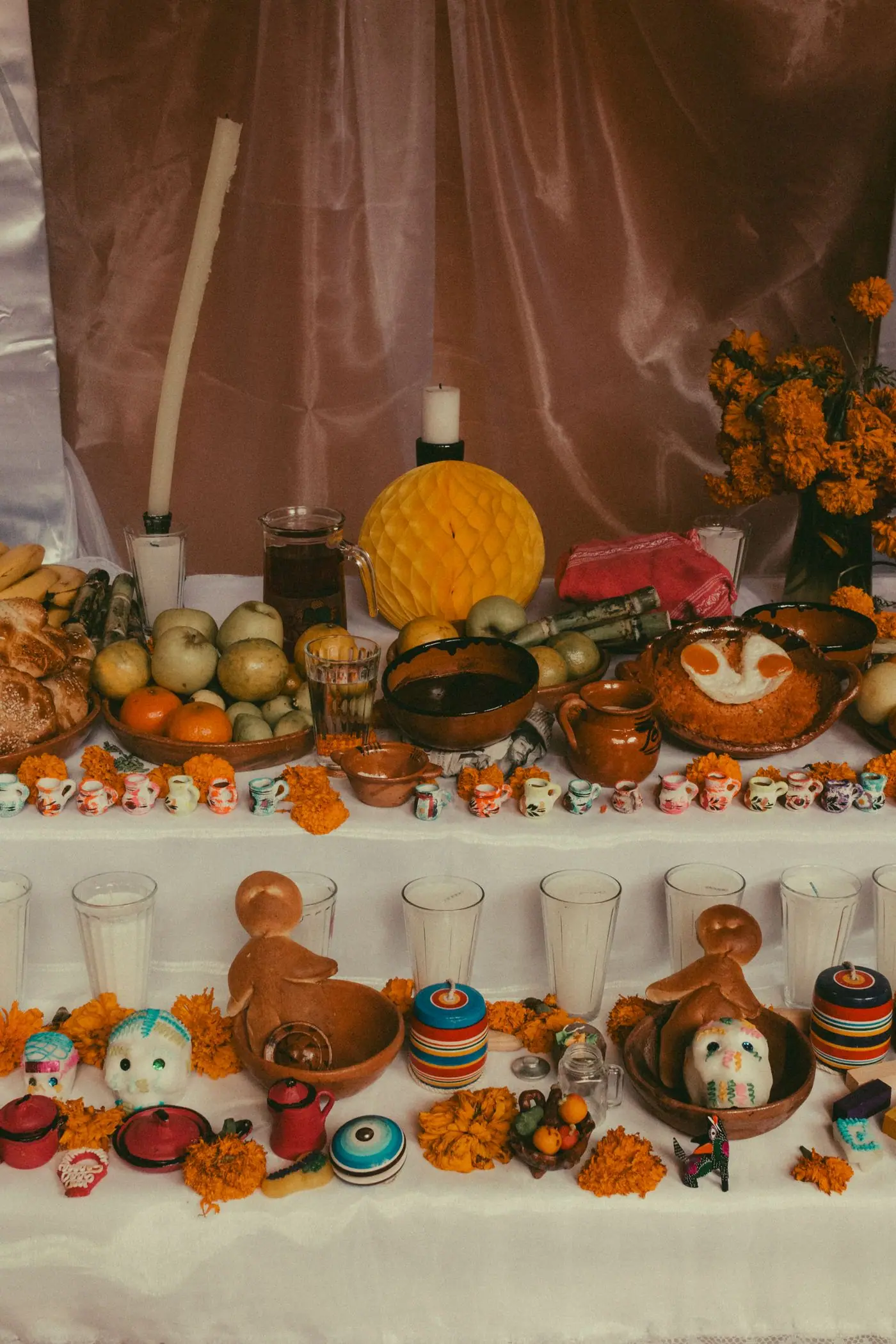 A full home ofrenda with pan de muerto, fruit, marigolds, sugar skulls, and rows of tiny clay mugs.
