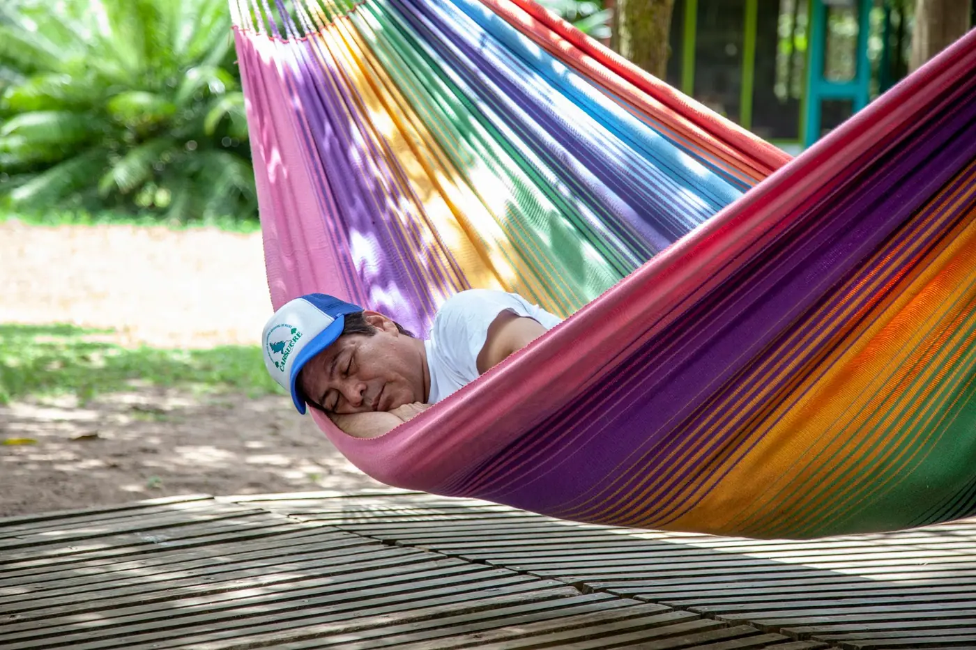 A man asleep in a striped hammock with his cap pulled down over his eyes