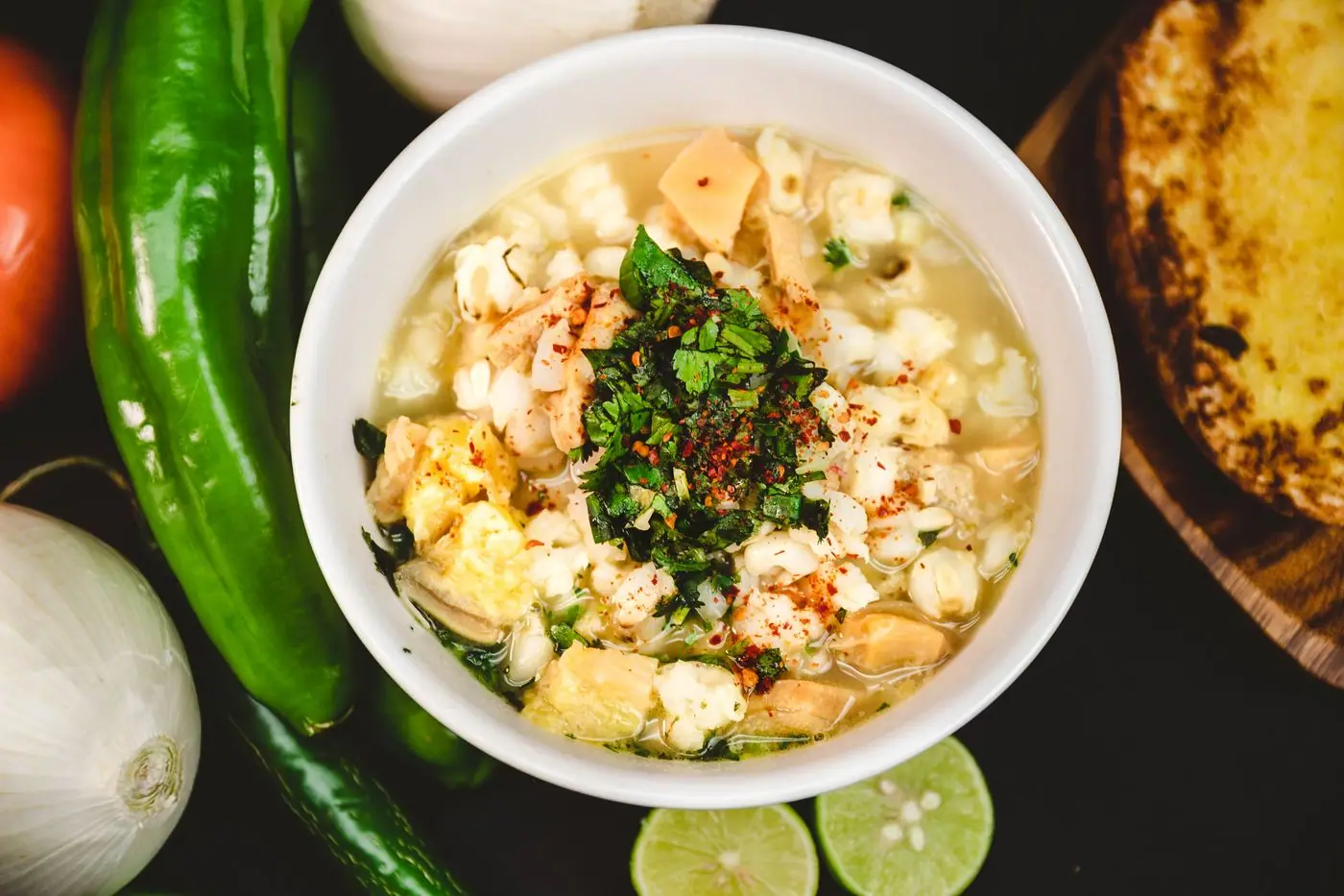 A bowl of pozole surrounded by small plates of radish, lettuce, onion, lime, and tostadas.