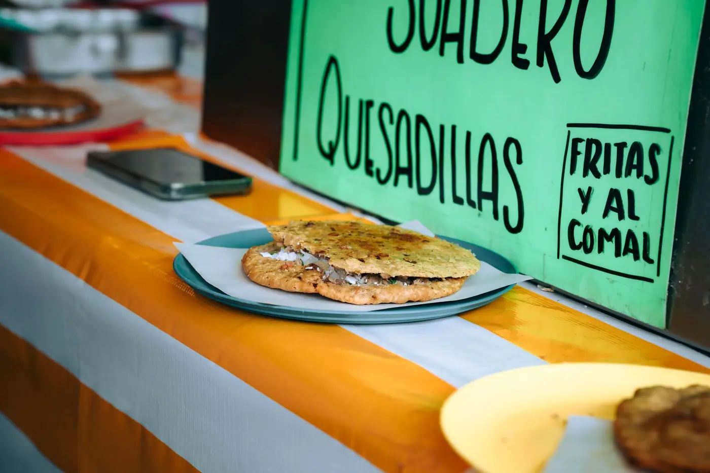 A quesadilla on a plate at a street stall, beside a hand-lettered sign reading suadero, quesadillas, fritas y al comal