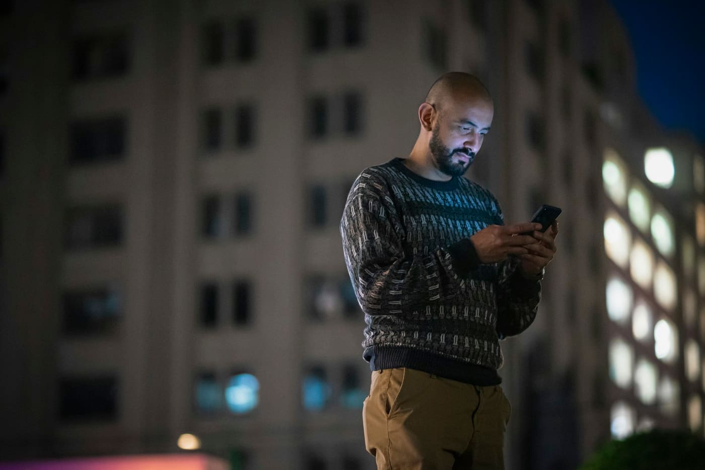 A man standing on a city street at night, lit by his phone screen, tall apartment building behind him.
