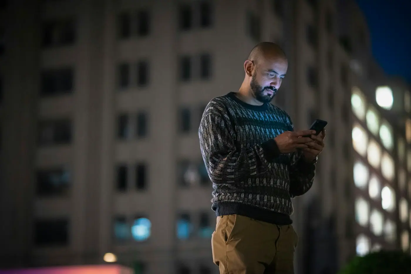 A man standing on a city street at night, lit by his phone screen, tall apartment building behind him.