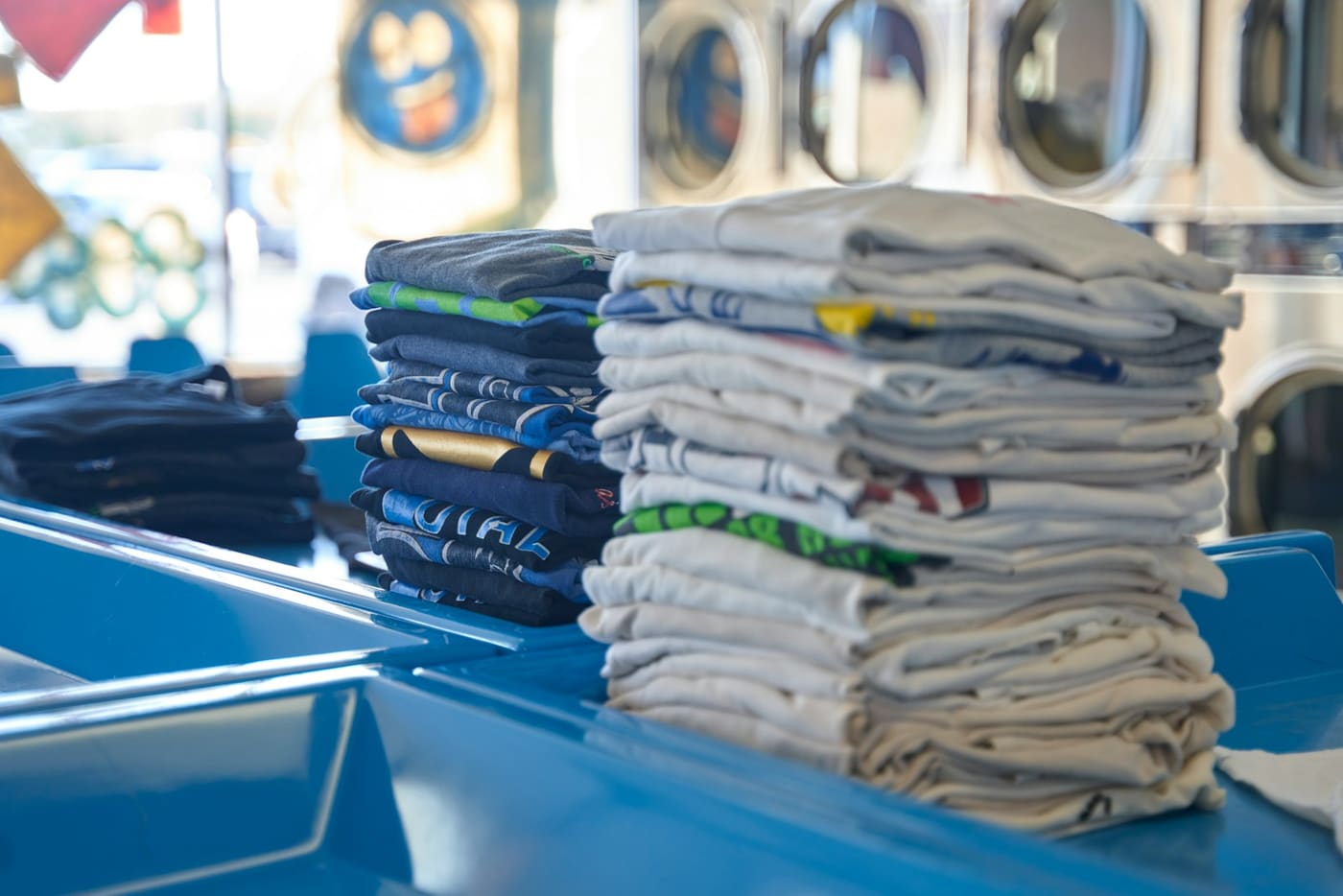 Neatly folded stacks of shirts and towels on a blue counter in a laundromat.