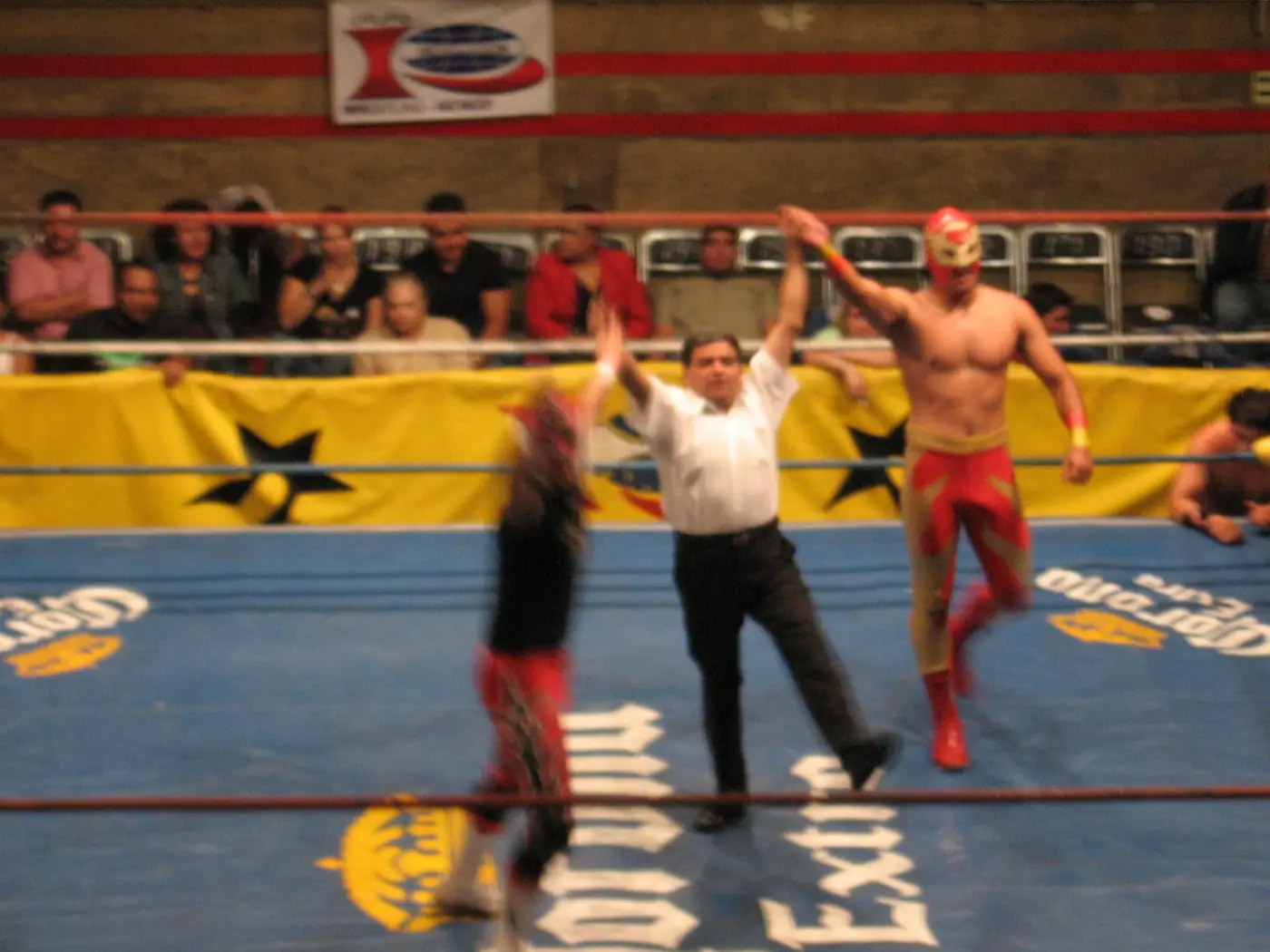A masked luchador raising his arms as the referee signals, crowd close behind