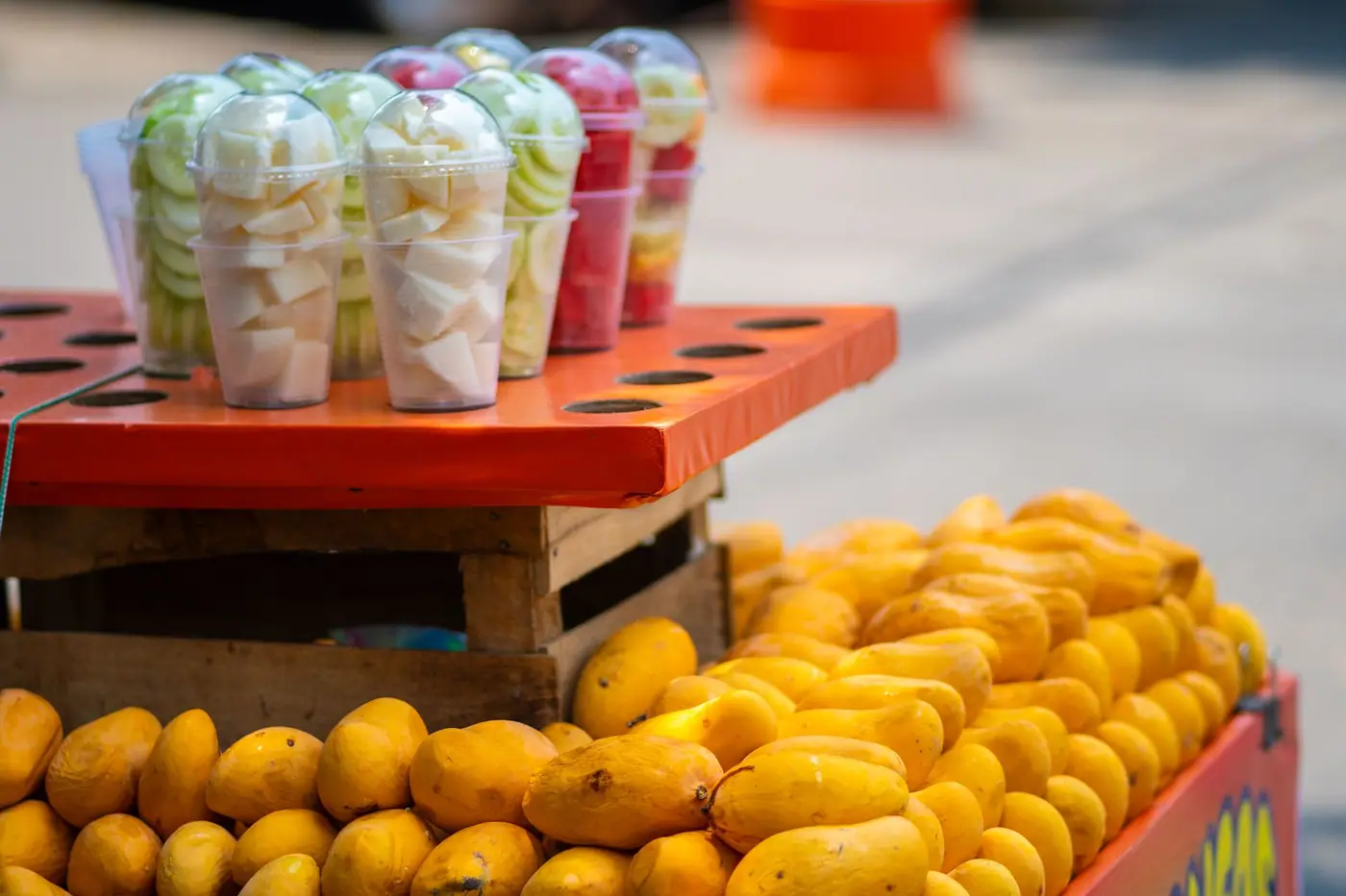 Cups of cut fruit stacked on a crate above a tray of mangoes at a street stall
