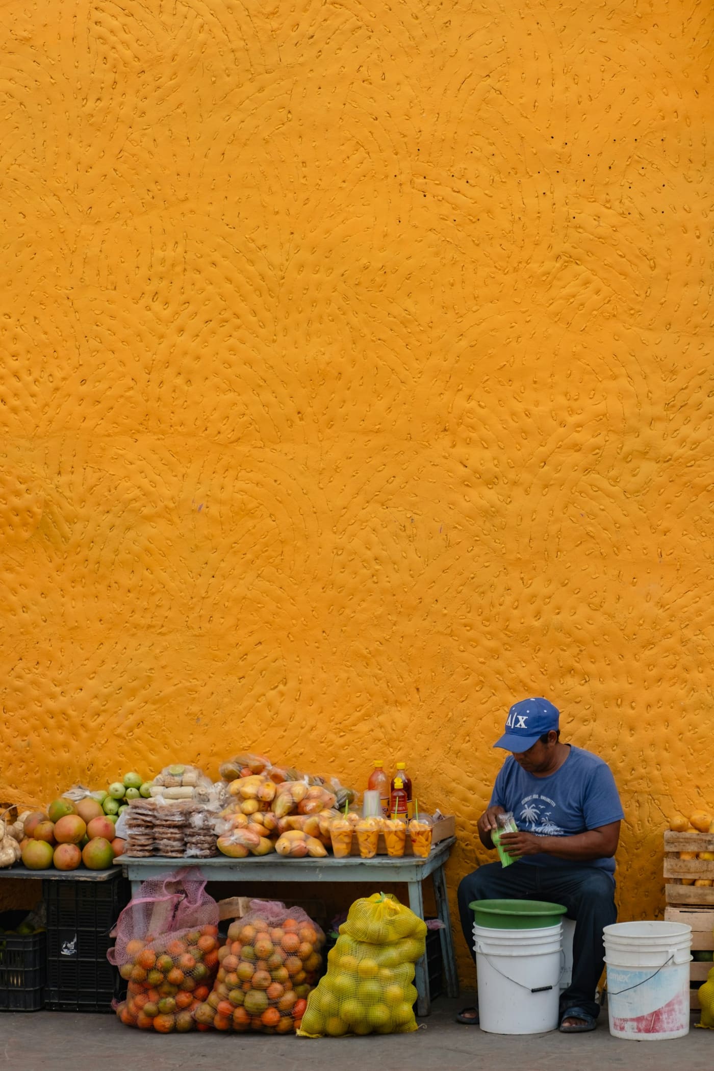 A man sitting beside a fruit stand in front of a textured orange wall.