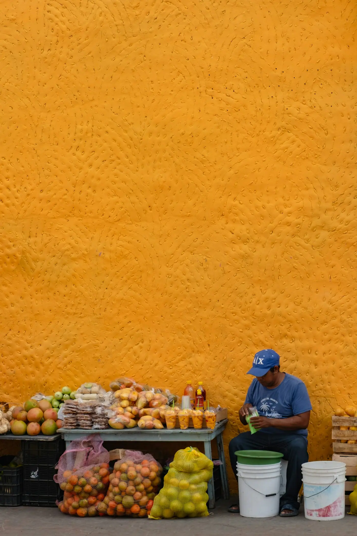 A man sitting beside a fruit stand in front of a textured orange wall.