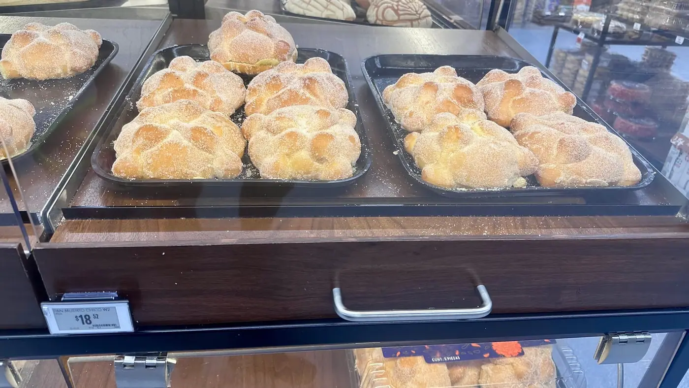 Trays of plain sugar-dusted pan de muerto in a supermarket bakery case, with a shelf label reading PAN MUERTO CHICO.