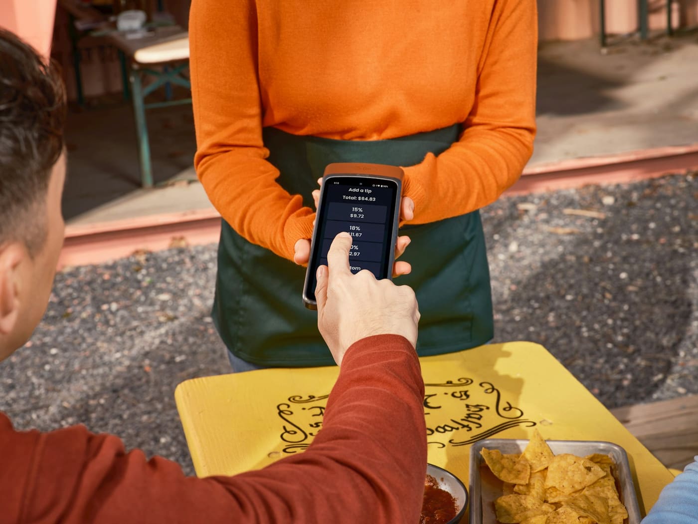 A person in an orange sweater taking a card payment on a phone at an outdoor table.