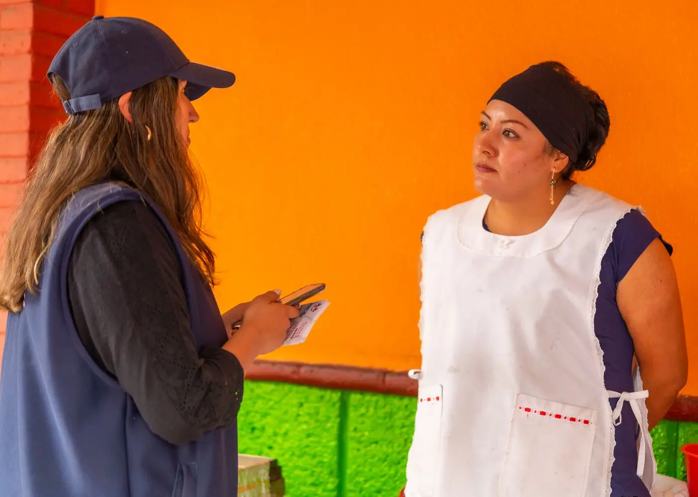 Two women standing and talking in front of a brightly painted wall in Mexico City.