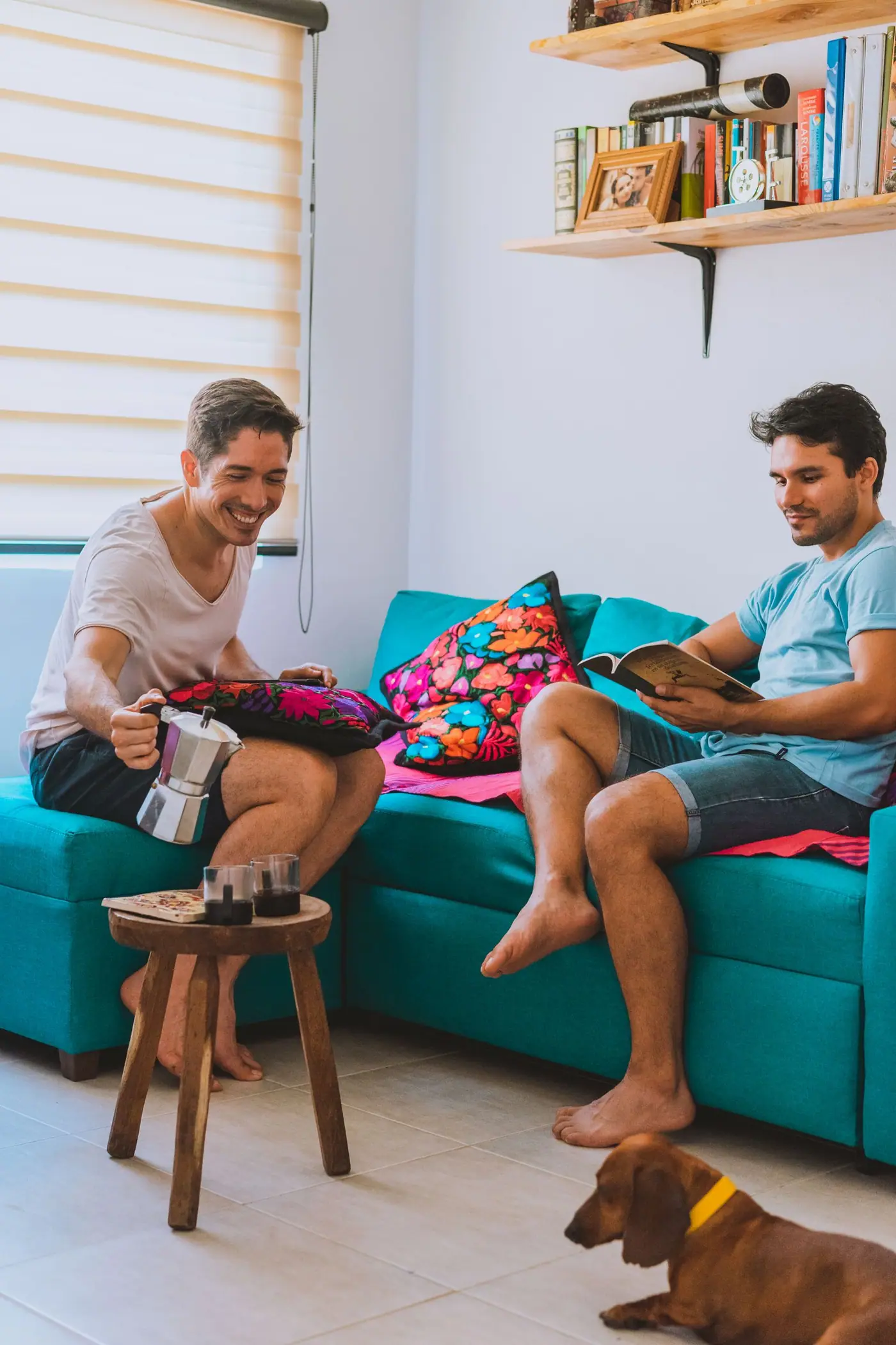 Two men drinking coffee on a teal couch in a living room with embroidered cushions.