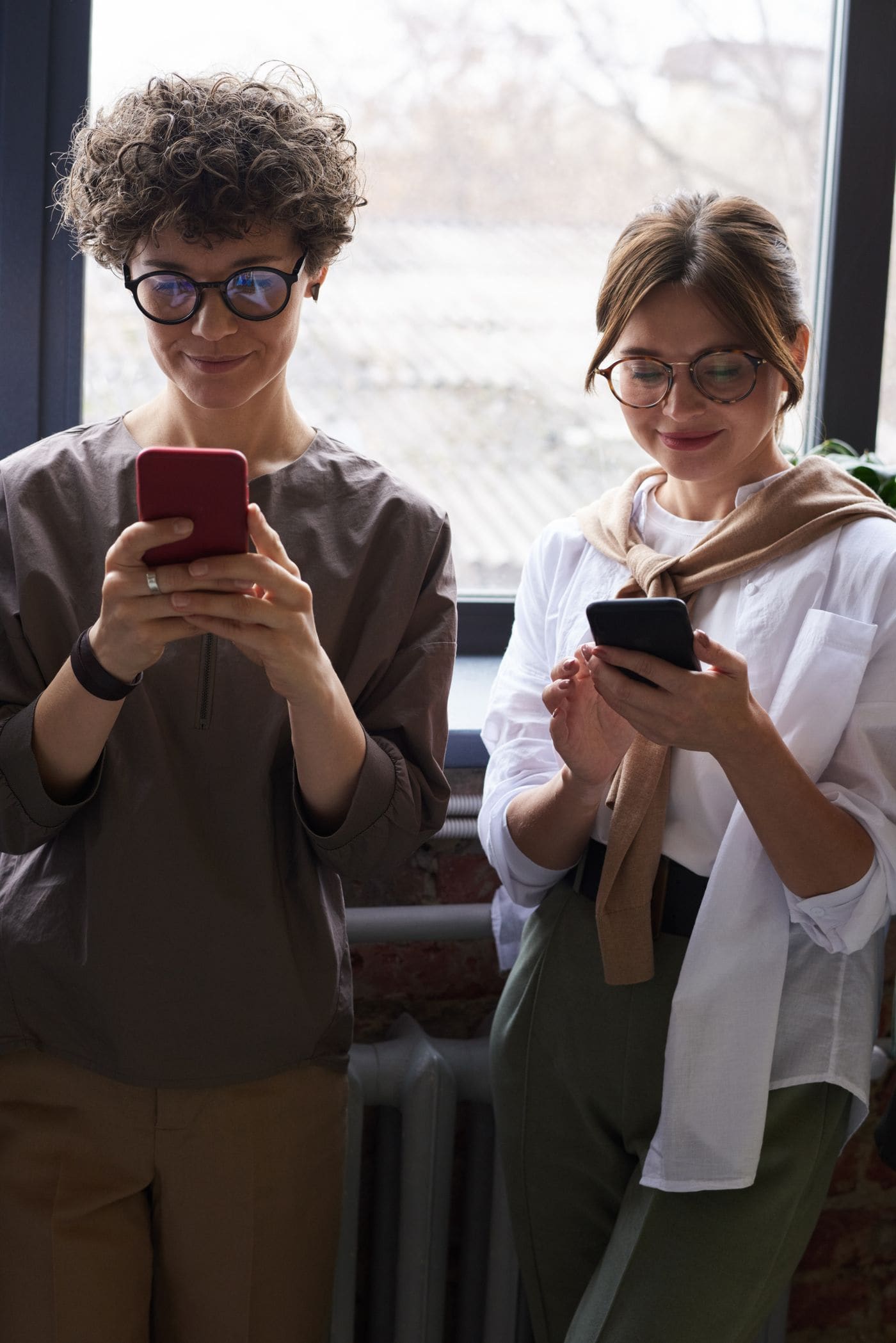 Two friends smiling while each looks at a smartphone indoors.