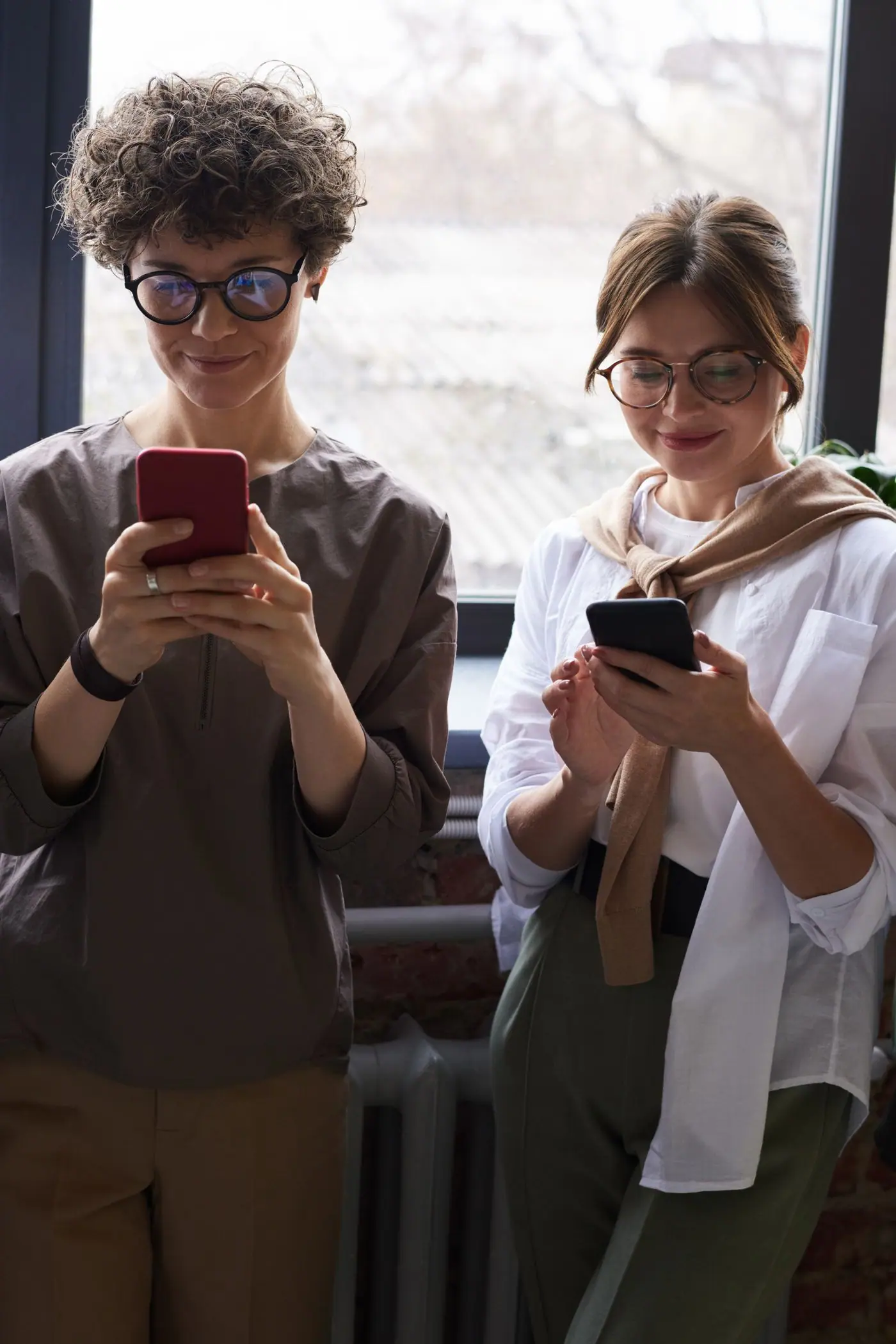 Two friends smiling while each looks at a smartphone indoors.