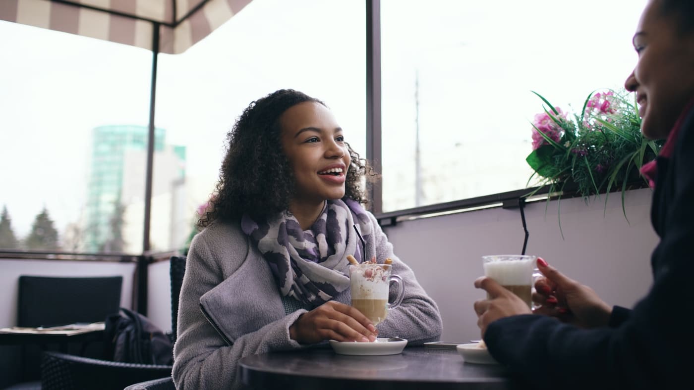 Two people talking and laughing over drinks at a cafe table by a window.