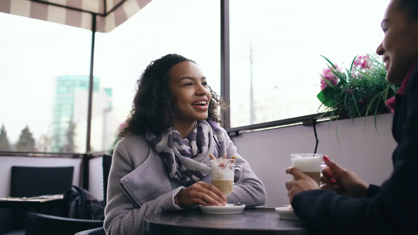 Two people talking and laughing over drinks at a cafe table by a window.