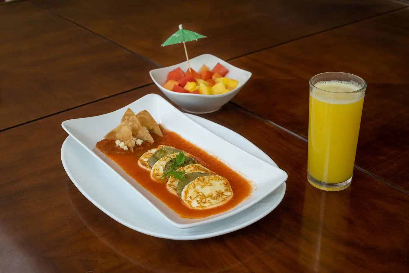A plate of eggs in salsa beside a small bowl of cut fruit and a glass of orange juice on a wooden table