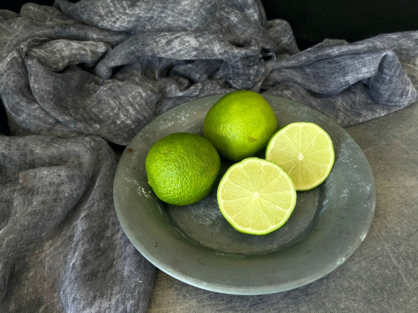 Whole and halved limes on a glass plate resting on grey cloth.