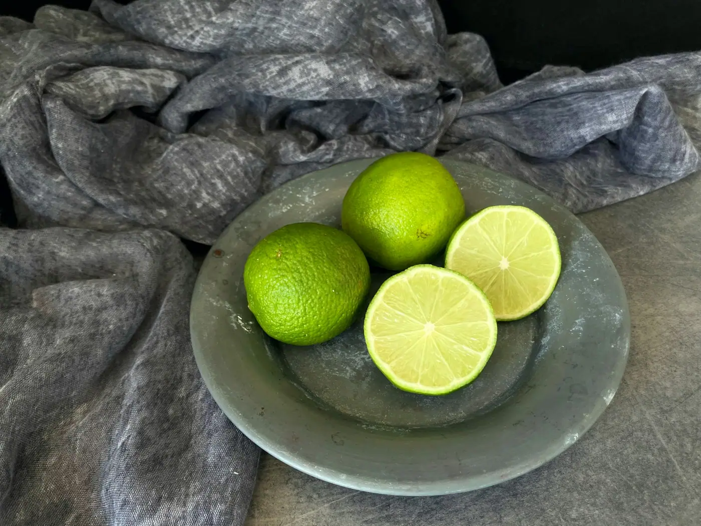 Whole and halved limes on a glass plate resting on grey cloth.