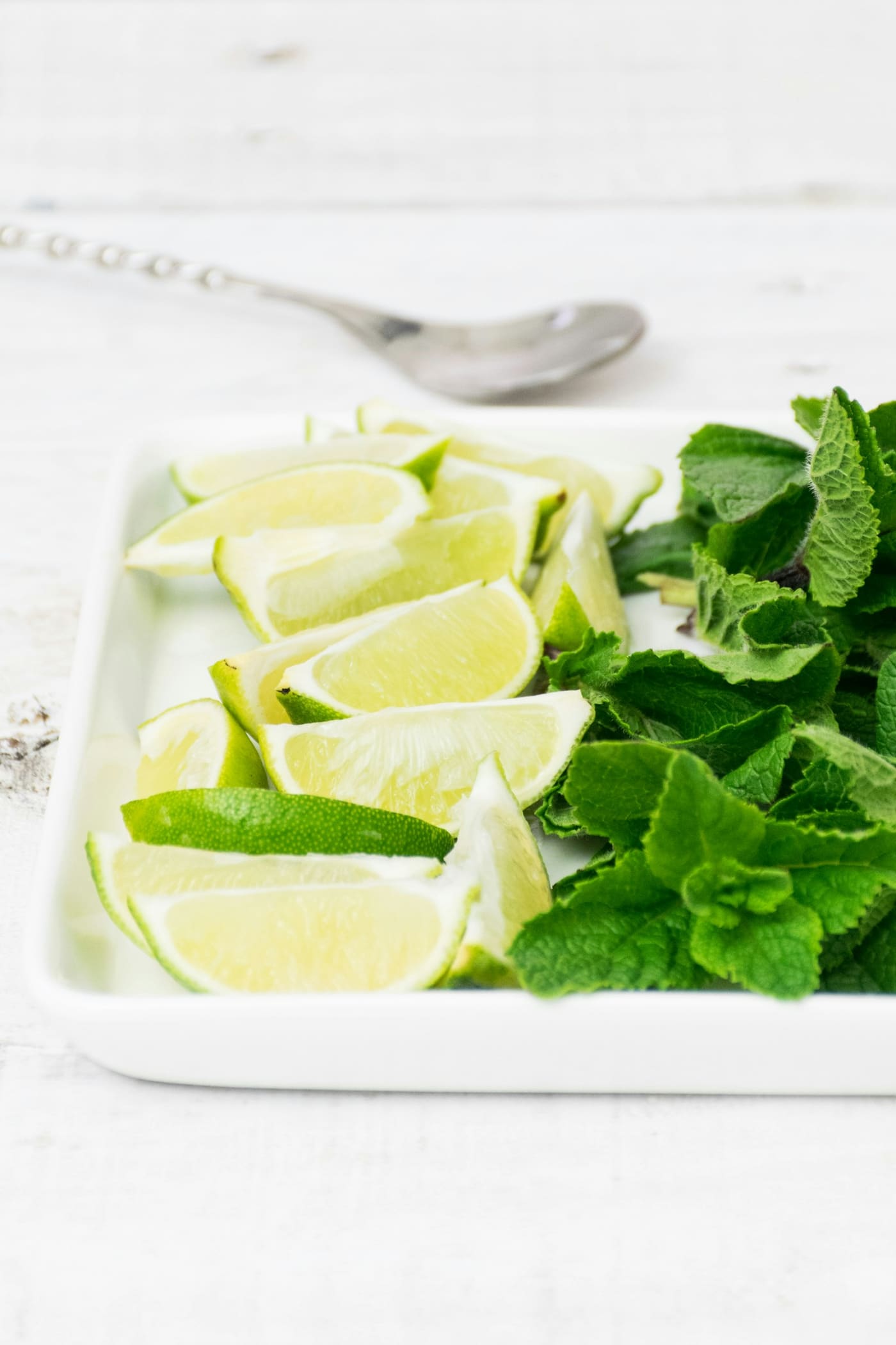Lime wedges and fresh green herb leaves arranged on a white tray with a spoon.