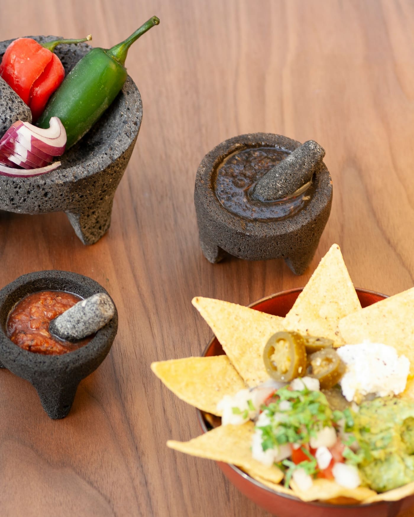 Stone molcajetes of salsa beside tortilla chips, guacamole and fresh chiles on a wooden table.
