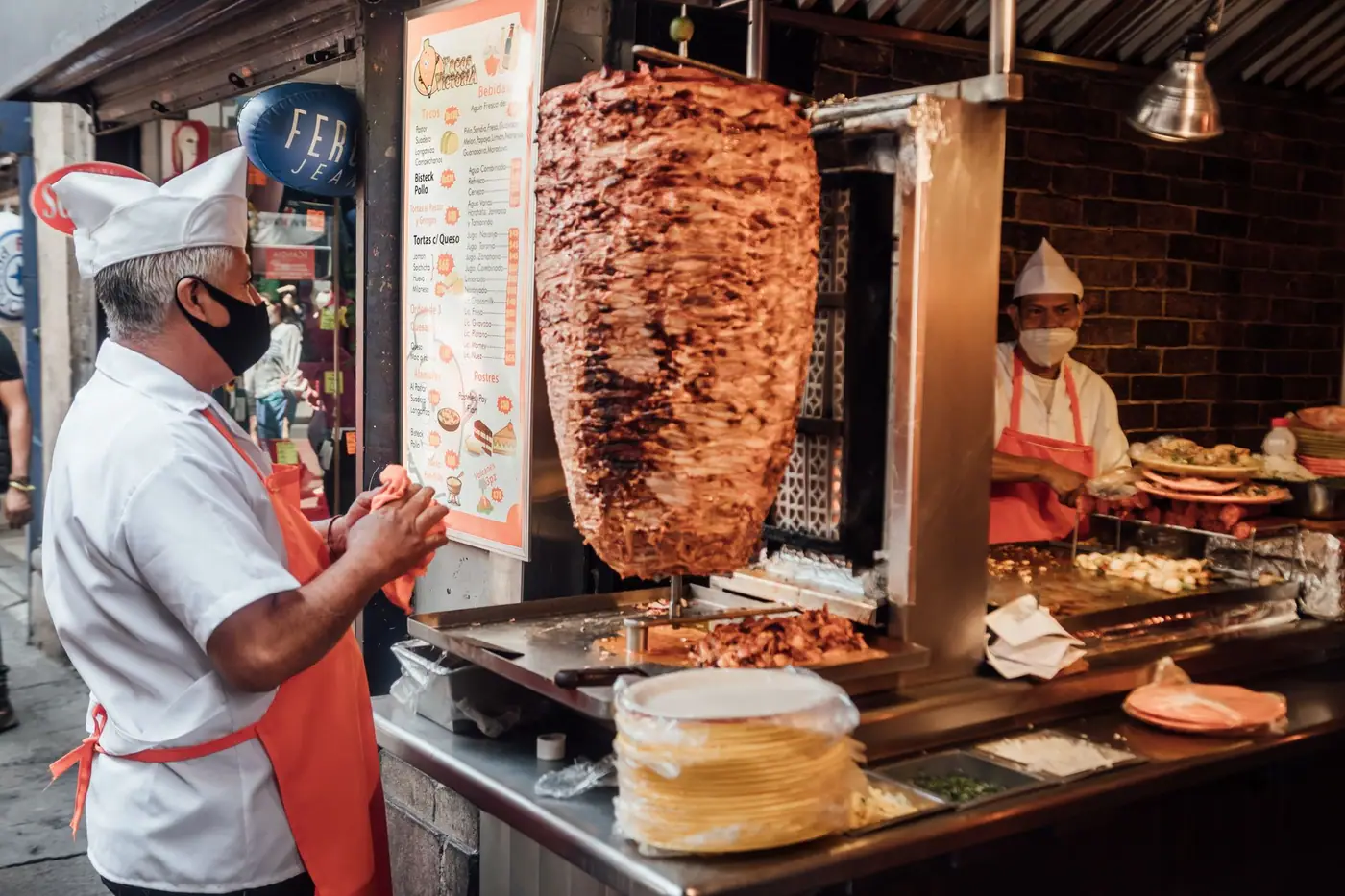 Two taqueros working a trompo of al pastor behind a lit counter, one shaving meat off the spit