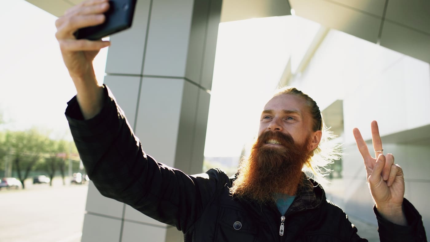 A bearded man holding a phone up to take a selfie outdoors, making a peace sign.
