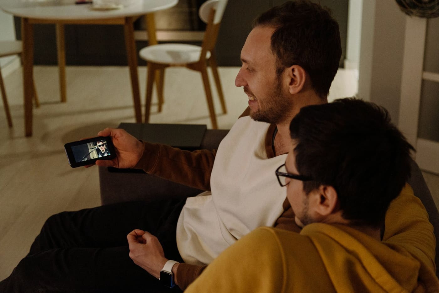Two men sitting close together on a sofa, both looking at the same phone screen.