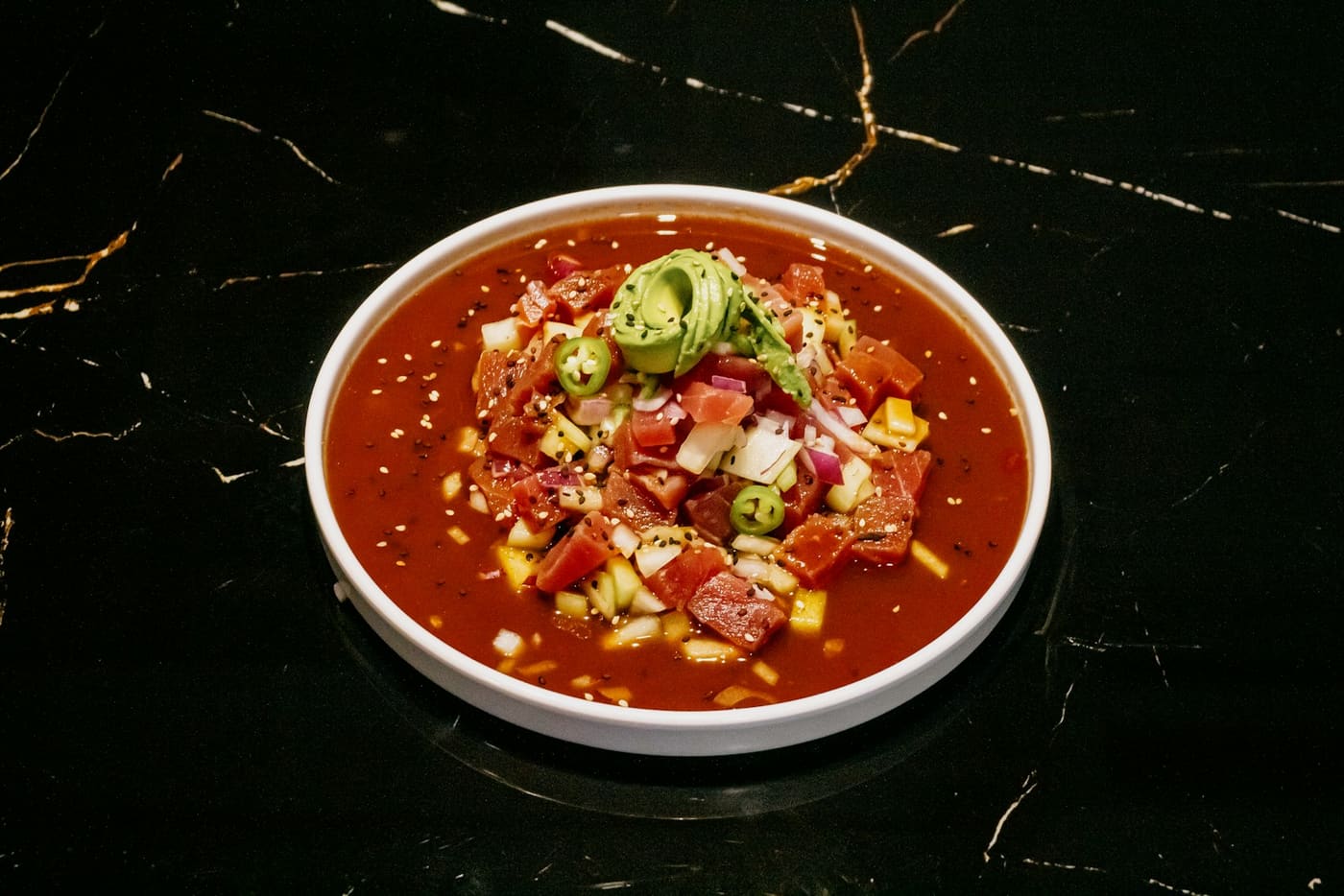 A white bowl of red tomato-based sauce topped with avocado and chopped chile.