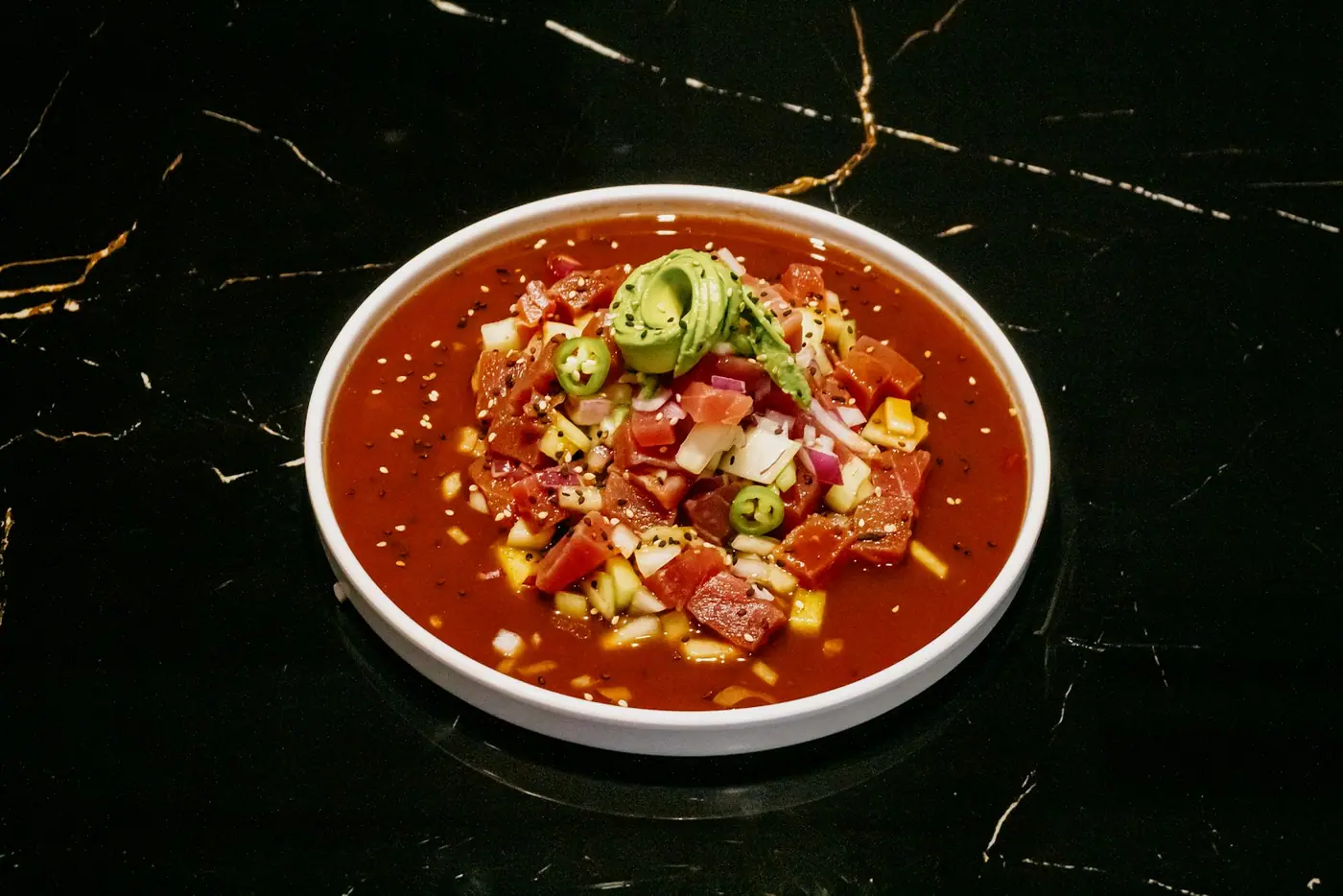A white bowl of red tomato-based sauce topped with avocado and chopped chile.
