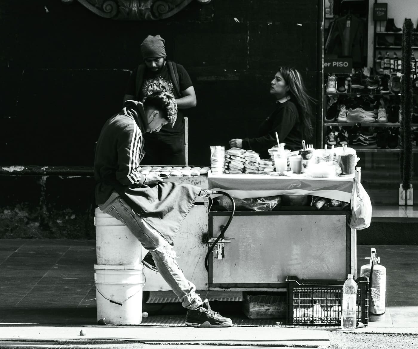 Black and white photo of a Mexican street food cart with people gathered around it on the sidewalk.