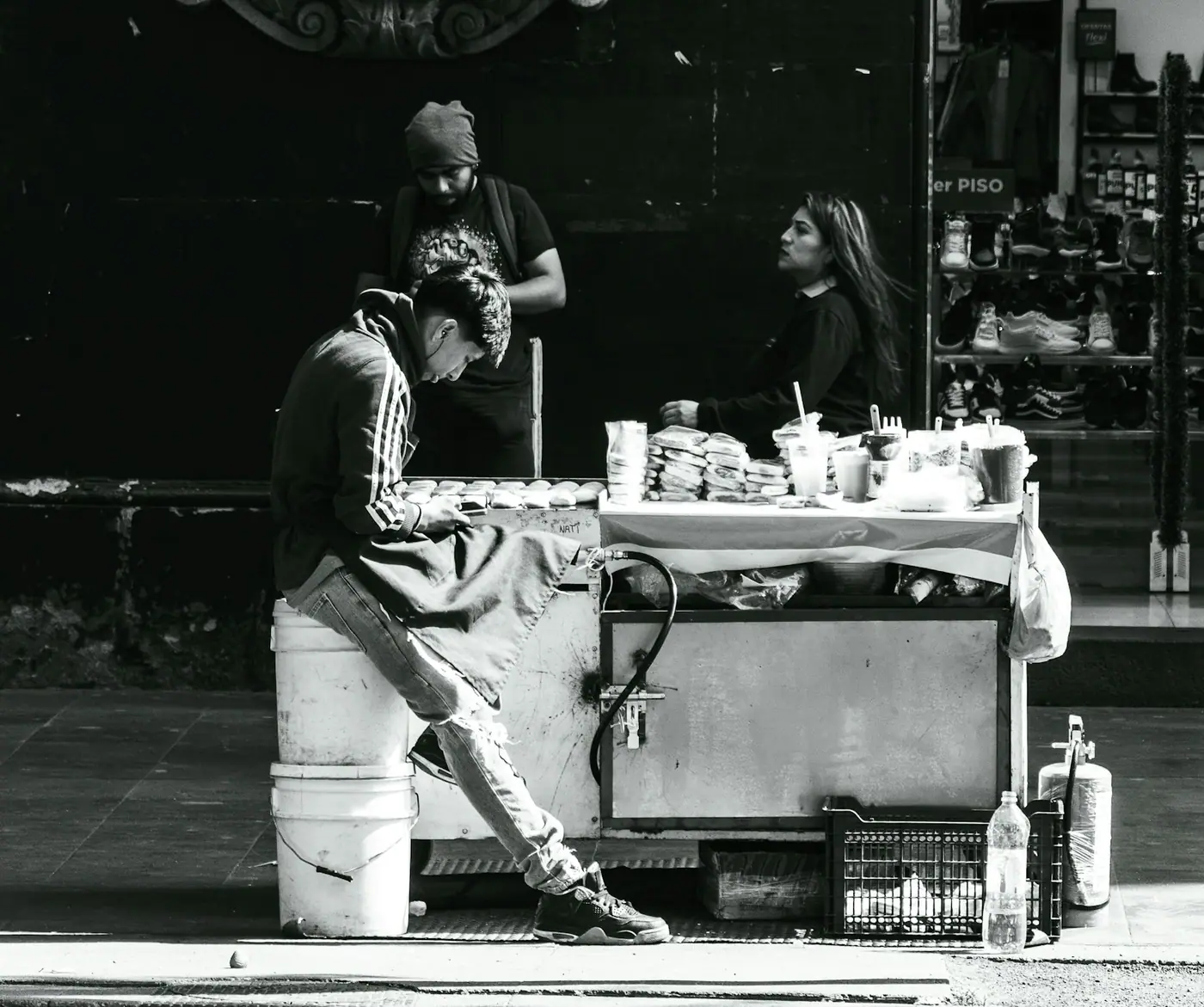 Black and white photo of a Mexican street food cart with people gathered around it on the sidewalk.