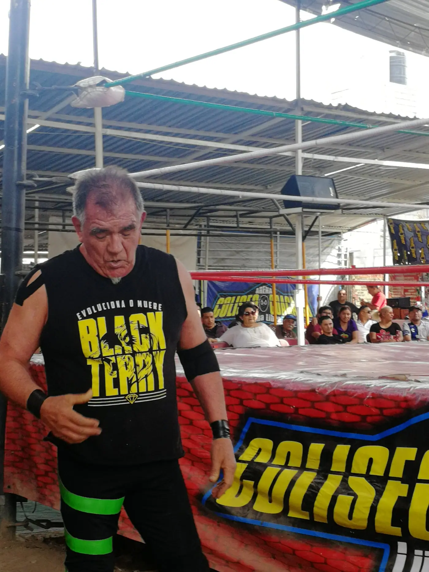 A veteran luchador standing at ringside in a small arena