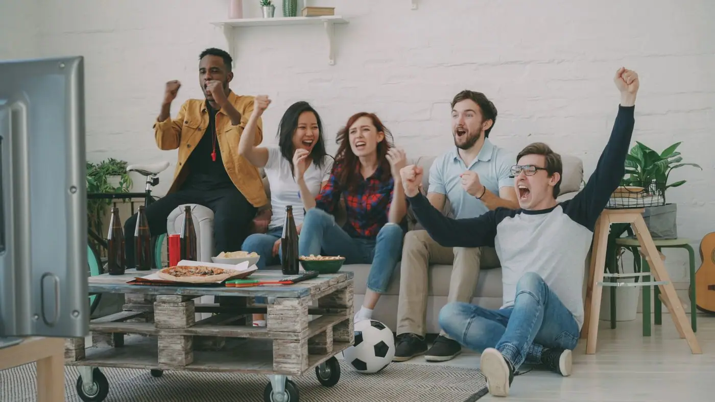 A living room full of friends reacting to a tense moment on the TV during a soccer match.