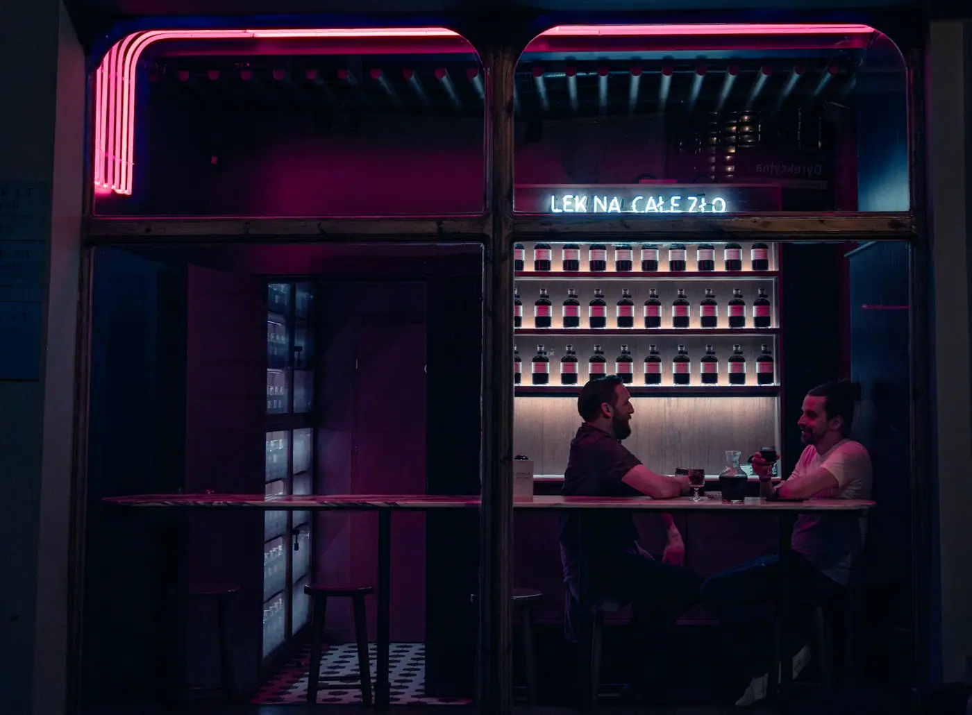 Two men talking over drinks at a bar lit with pink neon.