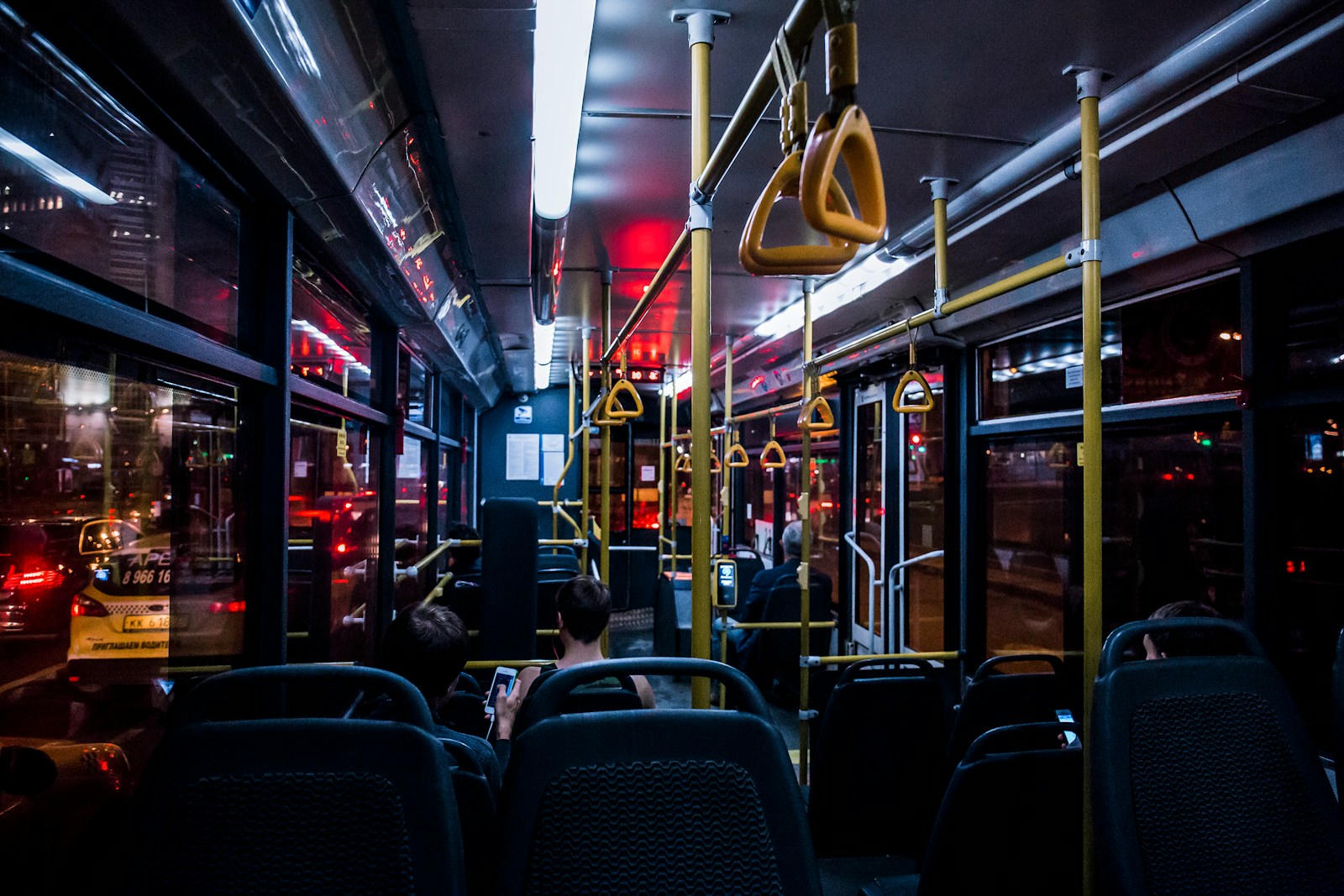 The interior of a bus at night, with metal handrails, overhead grab bars, and rows of empty seats.
