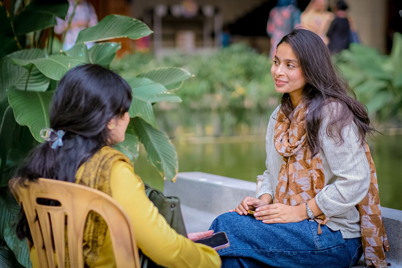Two women talking beside a green garden pond.
