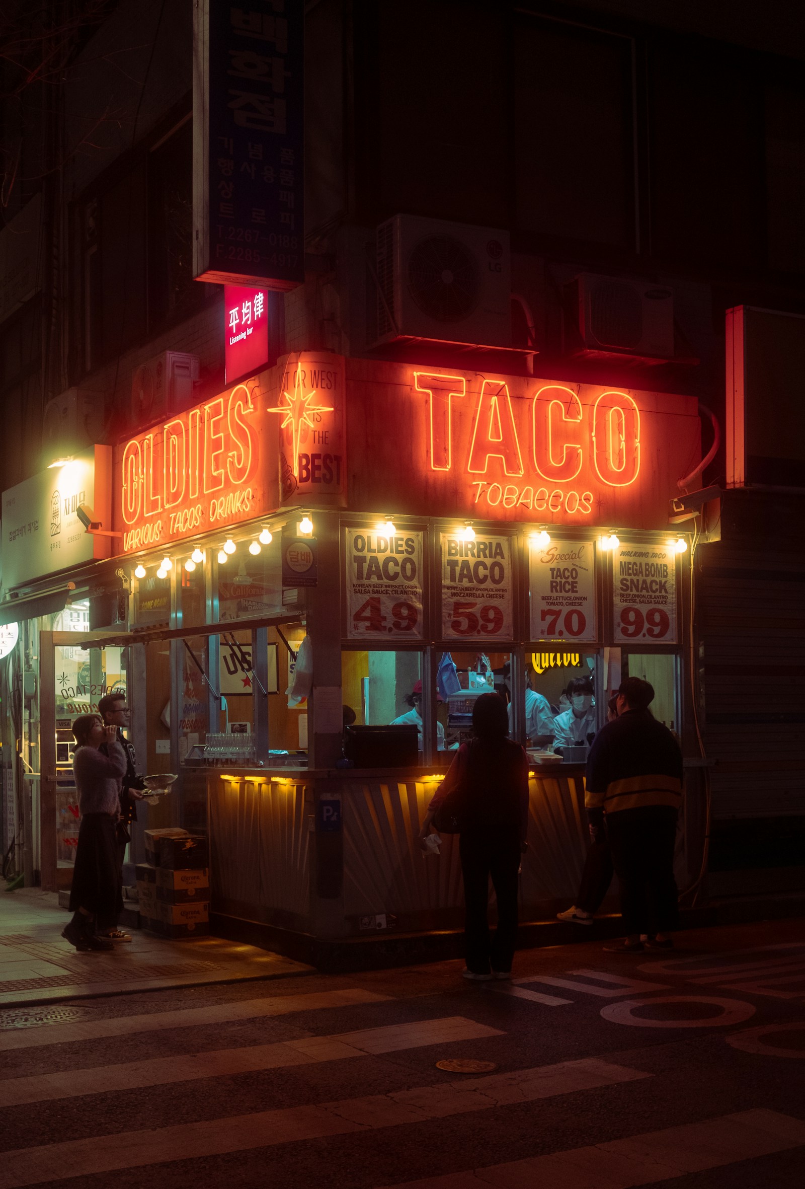 A neon-lit taco spot on a street corner at night.