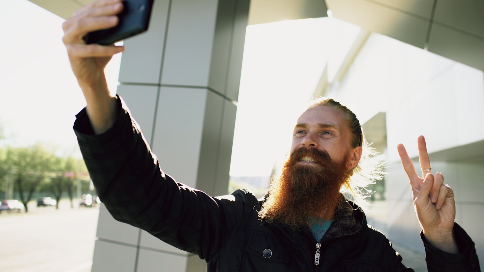 A bearded man holding a phone up to take a selfie outdoors, making a peace sign.