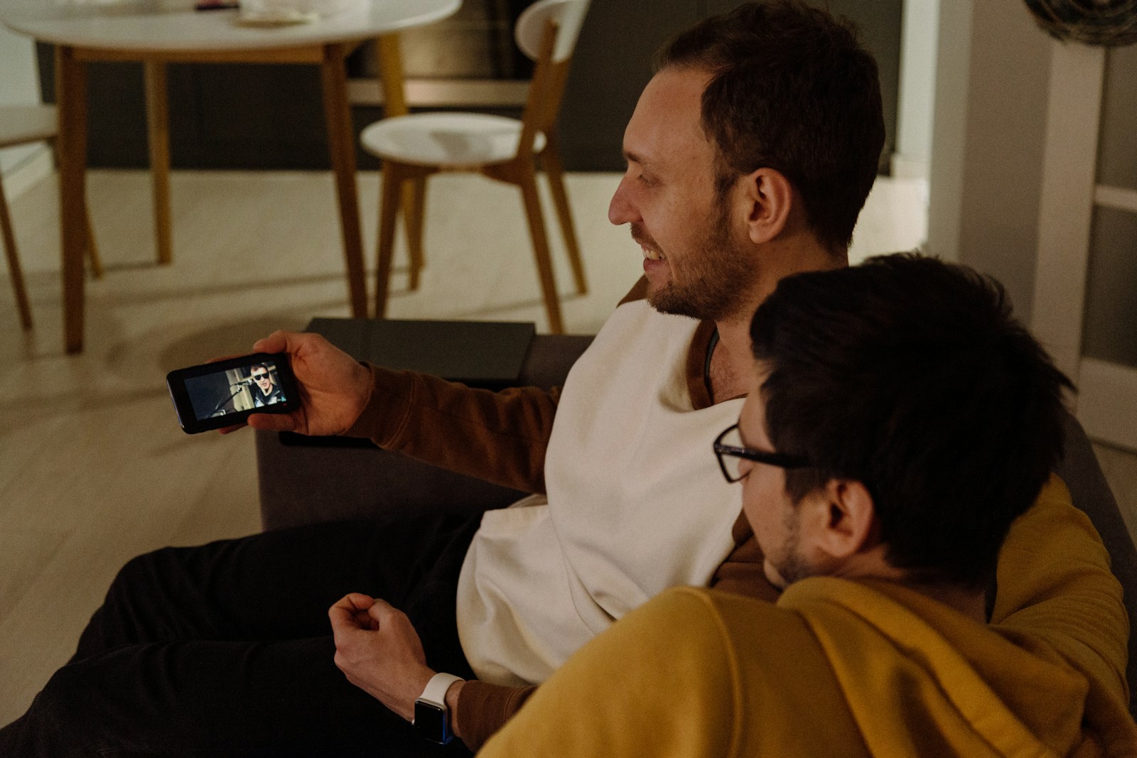 Two men sitting close together on a sofa, both looking at the same phone screen.