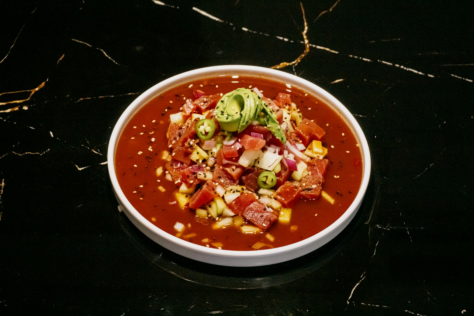 A white bowl of red tomato-based sauce topped with avocado and chopped chile.