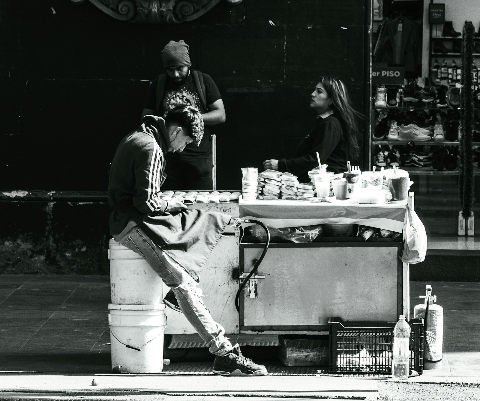 Black and white photo of a Mexican street food cart with people gathered around it on the sidewalk.