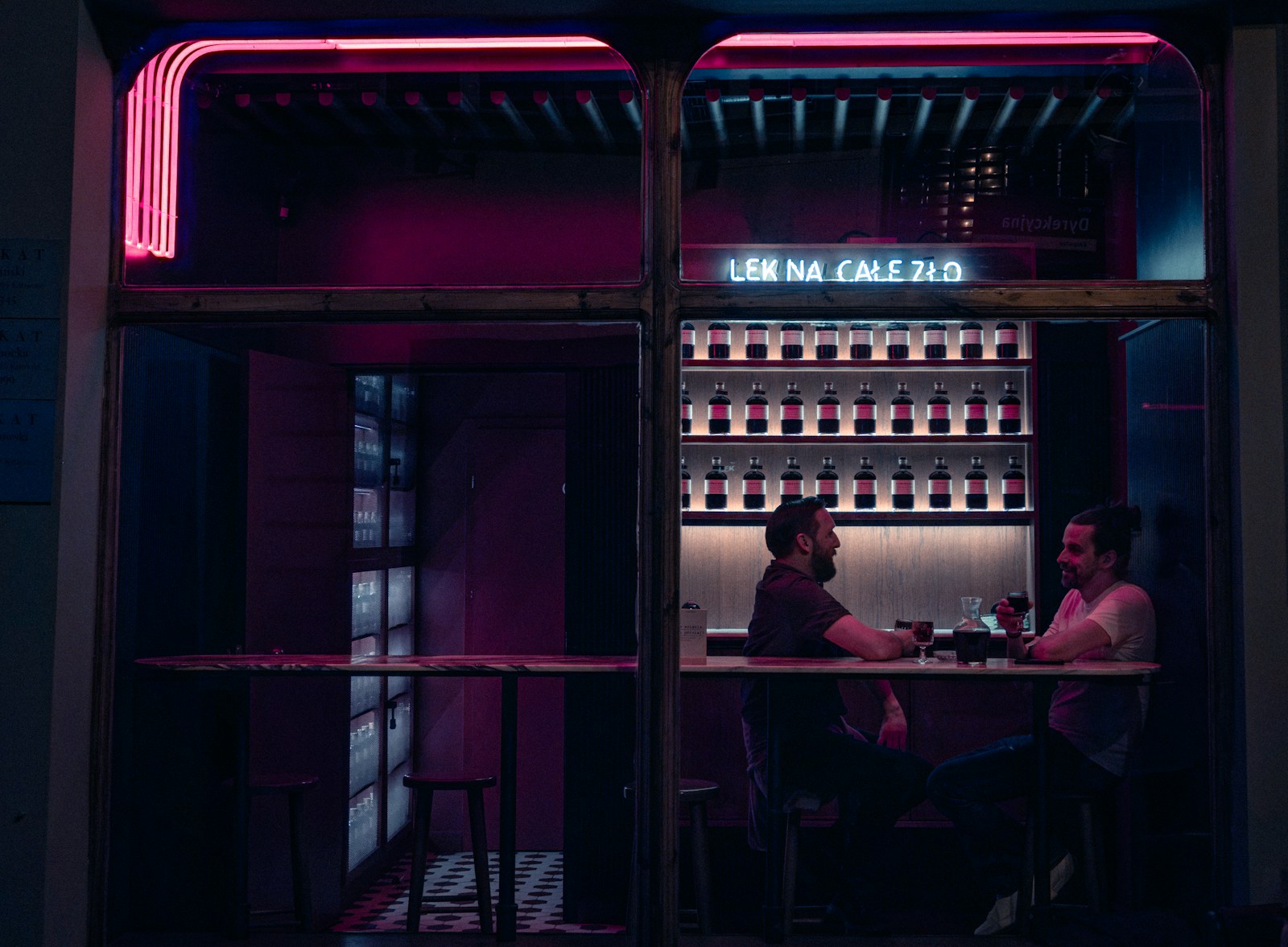 Two men talking over drinks at a bar lit with pink neon.